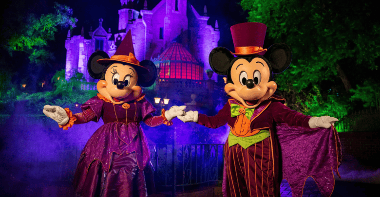 Mickey and Minnie Mouse are dressed in Halloween-themed costumes—Minnie in a purple witch outfit and hat, Mickey in a purple suit with an orange vest and green bow tie. They are holding hands and posing in front of a spooky, illuminated castle surrounded by mist.