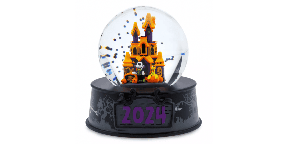 A Halloween-themed snow globe featuring a black and orange haunted house with bats inside the globe. The black base has tree silhouettes and a purple "2024" label on the front. Small Halloween decorations, including pumpkins, are placed near the house.