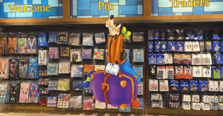Goofy sitting on a large, colorful pin in front of a wall filled with various collectible pins. The wall has sections labeled "Welcome," "Pin," and "Traders," showcasing a diverse collection.