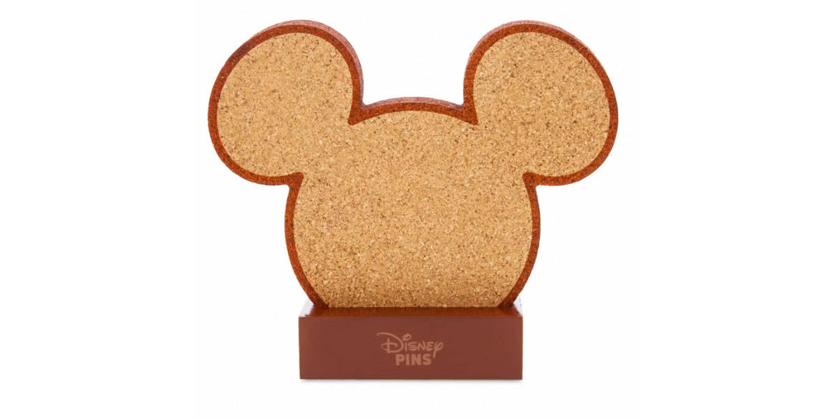 A cork board shaped like Mickey Mouse's head, set on a rectangular base with the words "Disney Pins" on it. The board has large circular ears and a round head, designed for pin display.