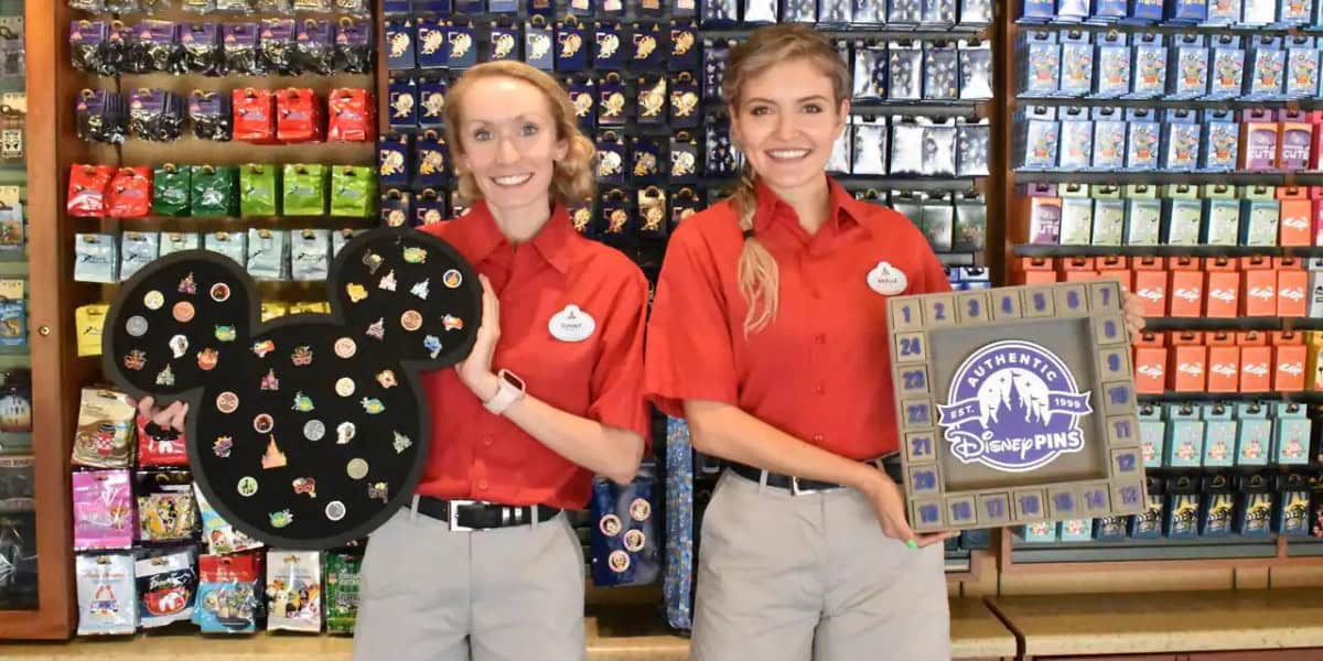 Two employees in red shirts and name badges stand in front of a display of colorful items. One holds a Mickey Mouse-shaped board with pins; the other holds a framed display featuring "Disney Pins." Shelves of merchandise are visible behind them.