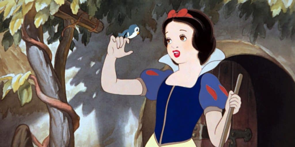 An animated woman with short black hair, dressed in a blue, red, and yellow dress, holds a broom and looks at a small blue bird perched on her hand—reminiscent of scenes from Disney movie remakes—standing outside a cottage under leafy branches.