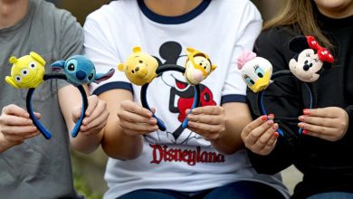 Three people holding Disney-themed headbands with plush characters: Alien, Stitch, Winnie the Pooh, Tigger, Daisy Duck, and Minnie Mouse. The person in the center wears a white Disneyland shirt.