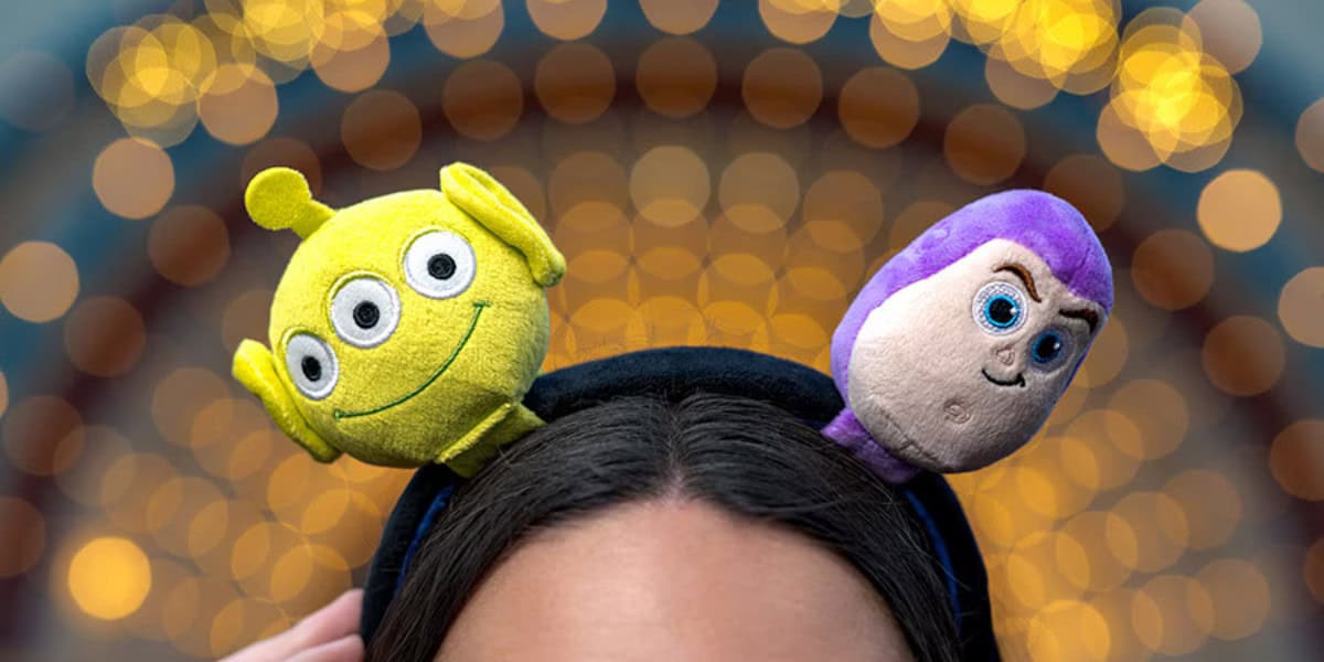 A person wearing a headband with plush toys of a green three-eyed alien and a purple-suited space ranger, with blurred decorative lights in the background.
