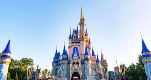 A large, ornate fairytale castle with blue and gold spires stands under a clear blue sky, surrounded by trees and flags.