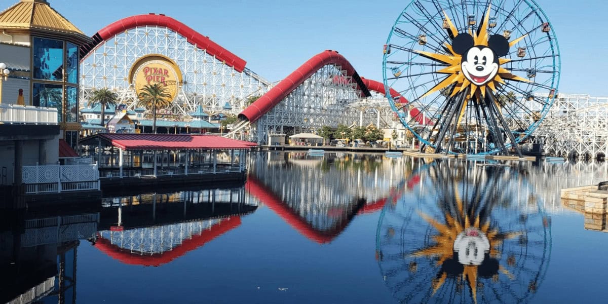 A calm lake reflects a red roller coaster and a large Ferris wheel with a smiling Mickey Mouse face at a sunny amusement park. Buildings and palm trees are visible along the water’s edge.