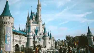 A large fairytale castle with blue spires stands under a bright sky, surrounded by visitors and trees, in a lively amusement park setting.