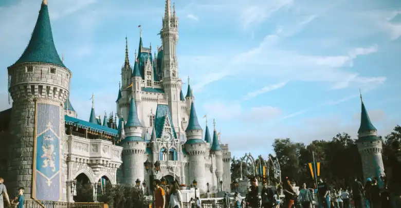 A large fairytale castle with blue spires stands under a bright sky, surrounded by visitors and trees, in a lively amusement park setting.