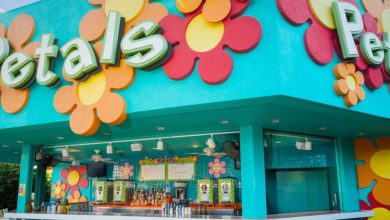 A colorful outdoor bar named "Petals" with large, bright flower decorations on a turquoise wall and a counter displaying bottles and drink machines. The menu is visible above the counter.