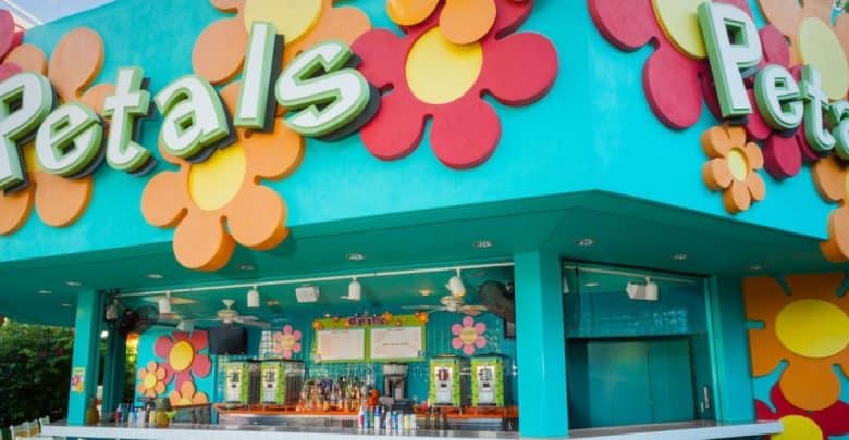 A colorful outdoor bar named "Petals" with large, bright flower decorations on a turquoise wall and a counter displaying bottles and drink machines. The menu is visible above the counter.