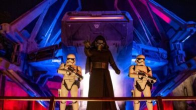 At Galaxy’s Edge, Kylo Ren stands in front of a Tie fighter, flanked by two First Order stormtroopers. The dramatic scene is enhanced by the red and blue lights casting an otherworldly glow.