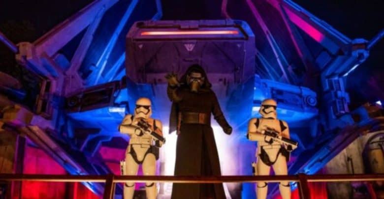 At Galaxy’s Edge, Kylo Ren stands in front of a Tie fighter, flanked by two First Order stormtroopers. The dramatic scene is enhanced by the red and blue lights casting an otherworldly glow.