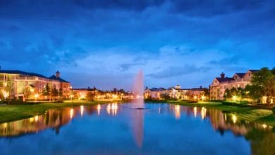 A serene lake with a central fountain is surrounded by well-lit buildings and trees at dusk, reflecting lights and a deep blue sky in the calm water.