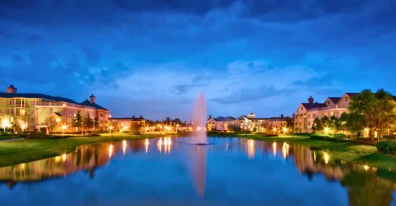 A serene lake with a central fountain is surrounded by well-lit buildings and trees at dusk, reflecting lights and a deep blue sky in the calm water.