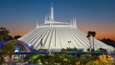 A futuristic white, conical building with spires, surrounded by greenery, is lit against a colorful sunset sky. This is Space Mountain, an amusement ride at a theme park.