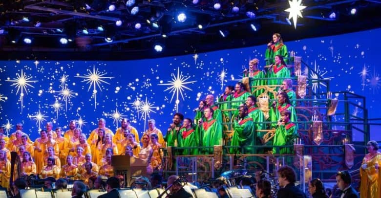 A large choir in yellow and green robes sings on a tiered stage decorated with bright star lights, backed by an orchestra. The backdrop is blue with glowing stars, creating a festive, celebratory atmosphere.
