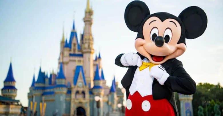 Mickey Mouse, wearing his classic outfit with red shorts, yellow bow tie, and white gloves, poses cheerfully in front of a blue and gold castle at a Disney theme park.