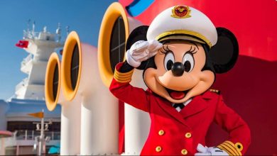 Minnie Mouse, dressed as a ship captain in a red uniform and white hat, stands and salutes in front of large yellow and red cruise ship funnels.