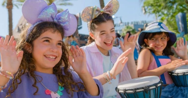 Three smiling children wearing colorful Disney mouse ear headbands and bracelets play drums and raise their hands outdoors on a sunny day, with palm trees and other people in the background.