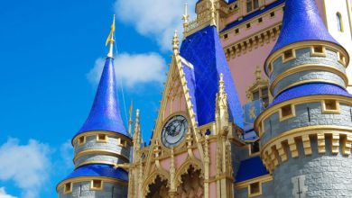 Close-up of a colorful fairytale castle in Magic Kingdom, with blue pointed roofs, gold accents, gray stone walls, and a clock above the entrance, set against a bright blue sky with scattered white clouds at Walt Disney World.
