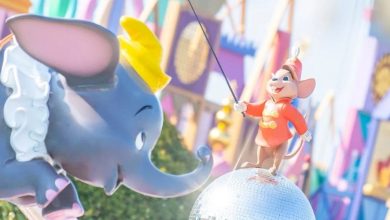 A Dumbo elephant statue with a yellow hat faces a smiling mouse figure in a red uniform standing on a reflective silver ball. Colorful decorations and geometric patterns are visible in the blurred background.
