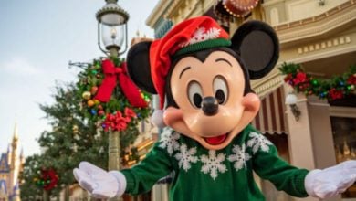 Mickey Mouse wearing a festive red Santa hat and green sweater with white snowflakes, standing outdoors on a decorated street with holiday garlands.