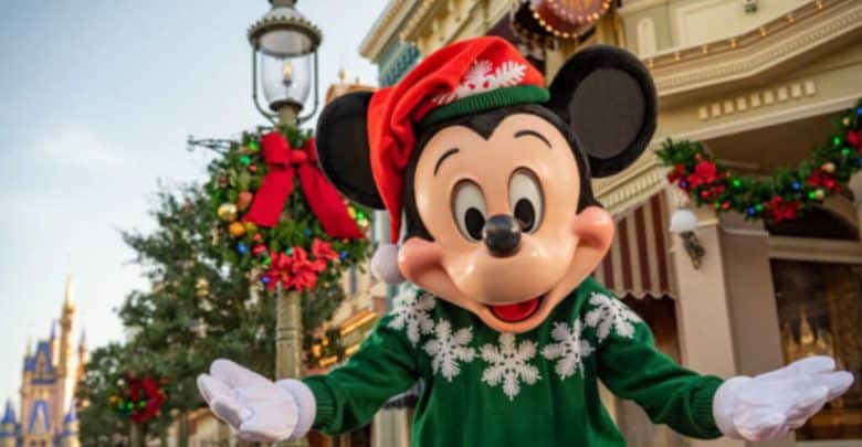 Mickey Mouse wearing a festive red Santa hat and green sweater with white snowflakes, standing outdoors on a decorated street with holiday garlands.