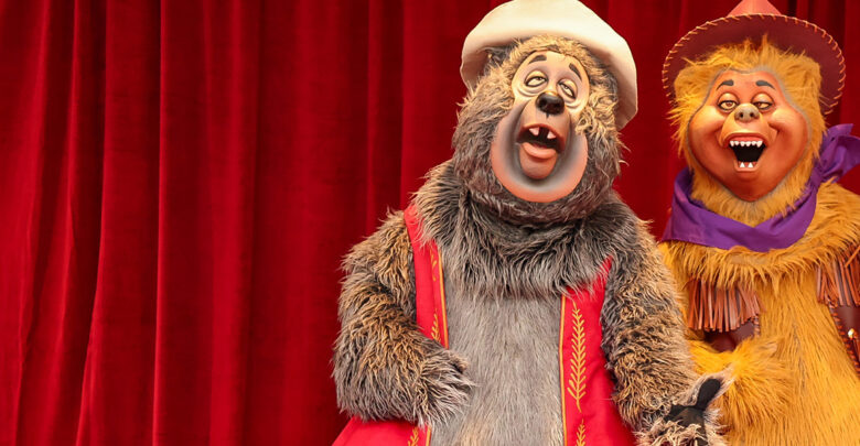 Big Al and Liver Lips McGrowl smile in their signature hats and vests before red curtains, capturing Frontierland’s whimsy.