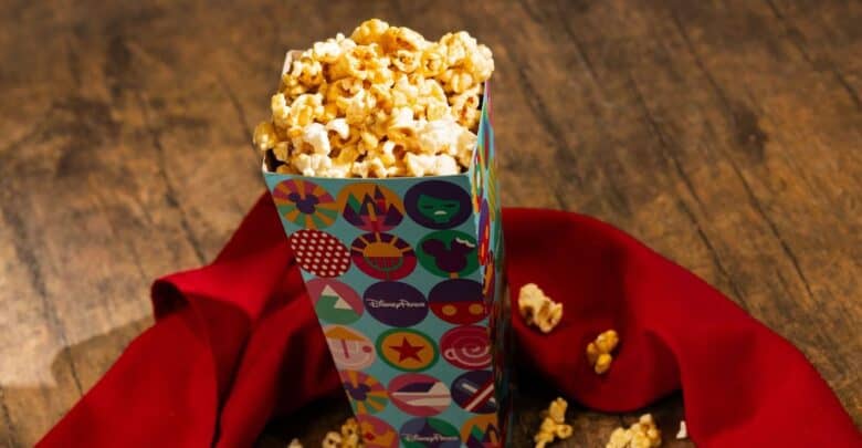 A festive Disney World popcorn box sits on red cloth, caramel popcorn spilling onto wood like a classic park snack experience.