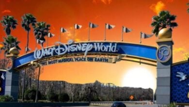 An emoji with a red face, sweat droplets, and its tongue sticking out is placed next to a fairytale castle under a bright blue sky with fluffy clouds. The image conveys a sense of heat and exhaustion, as if the character desperately needs heat relief in a magical theme park setting.