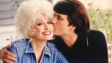 A couple shares a sweet moment at Dollywood, as a man kisses his smiling companion in her blonde hair and denim jacket, enjoying the lively atmosphere of the theme park’s outdoor setting.