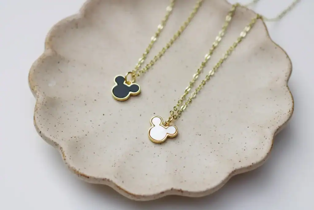 Cute Mickey Mouse charm necklaces on a beige ceramic dish, perfect Disney jewelry for fans.