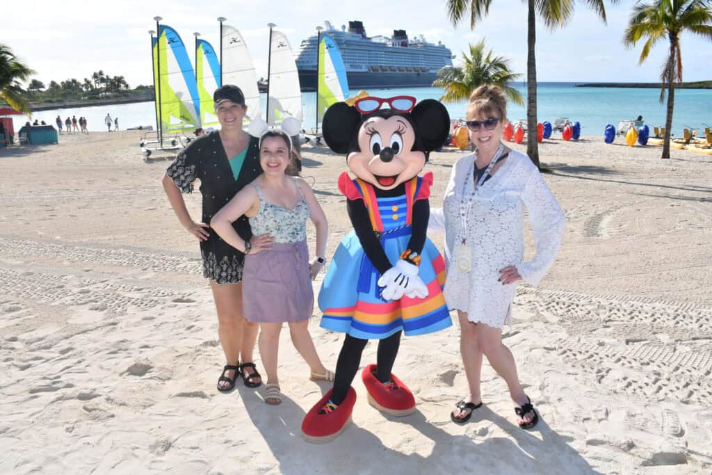 Girl and women with Minnie Mouse at tropical beach, sailboats, cruise ship in background, relaxing vacation scene.