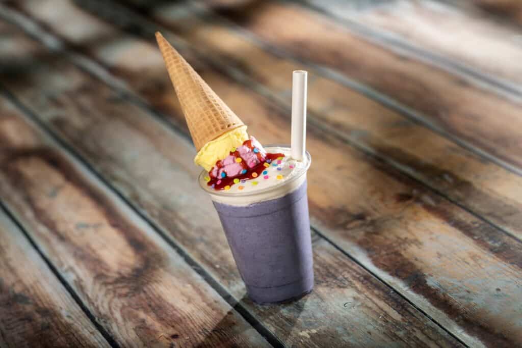Vibrant cookie dough milkshake topped with ice cream, sprinkles, and an upside-down wafer cone, on rustic wooden table.