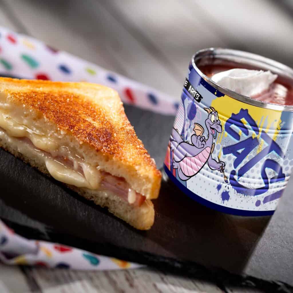 Colorful Disney dragons and characters on a hot chocolate mug with a grilled cheese sandwich.