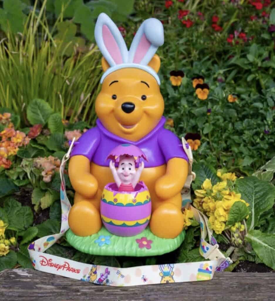 Easter Winnie the Pooh figurine wearing bunny ears, holding a decorated Easter egg, surrounded by co.