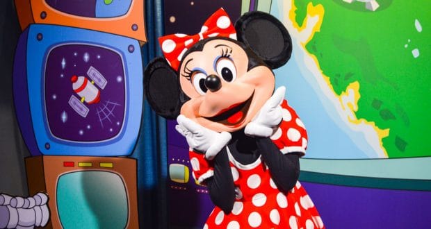 Minnie Mouse beams in her classic red polka-dot dress before vibrant cartoon screens and a globe at Walt Disney World.