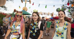 Three women wearing Toy Story-themed shirts, headbands, and green alien necklaces smile as they walk together through a lively, outdoor area at Hollywood Studios. Colorful lights, themed props, and umbrellas add to the fun—perfect for Hollywood Studios tips!.