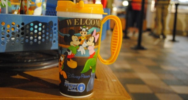 A Disney Parks refillable mug featuring beloved characters and "WELCOME" text rests on a wooden table inside the park.