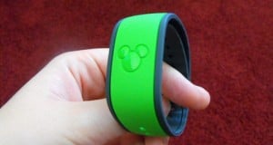 A guest displays a green Disney World FastPass+ wristband featuring the Mickey Mouse logo, with a vivid red backdrop.