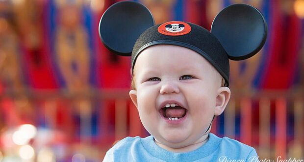 Smiling baby wearing Mickey Mouse ears hat, with an open mouth and visible teeth, in front of a colorful, blurred background.