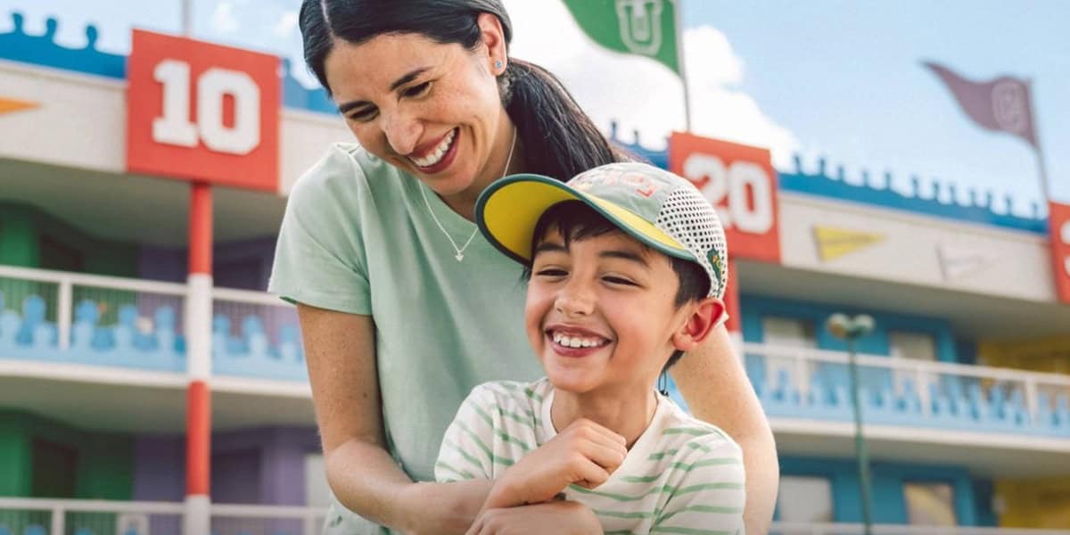 A smiling woman hugs a laughing young boy in a baseball cap outside a colorful building with numbered signs and flags at Hollywood Studios. The clear blue sky adds to their cheerful day—perfect for discovering new Hollywood Studios tips together.
