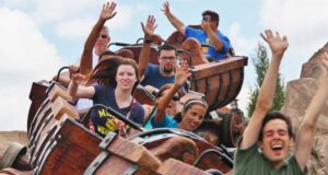 A group of people ride a wooden roller coaster, smiling and raising their hands in excitement as the train moves along the track under a partly cloudy sky.