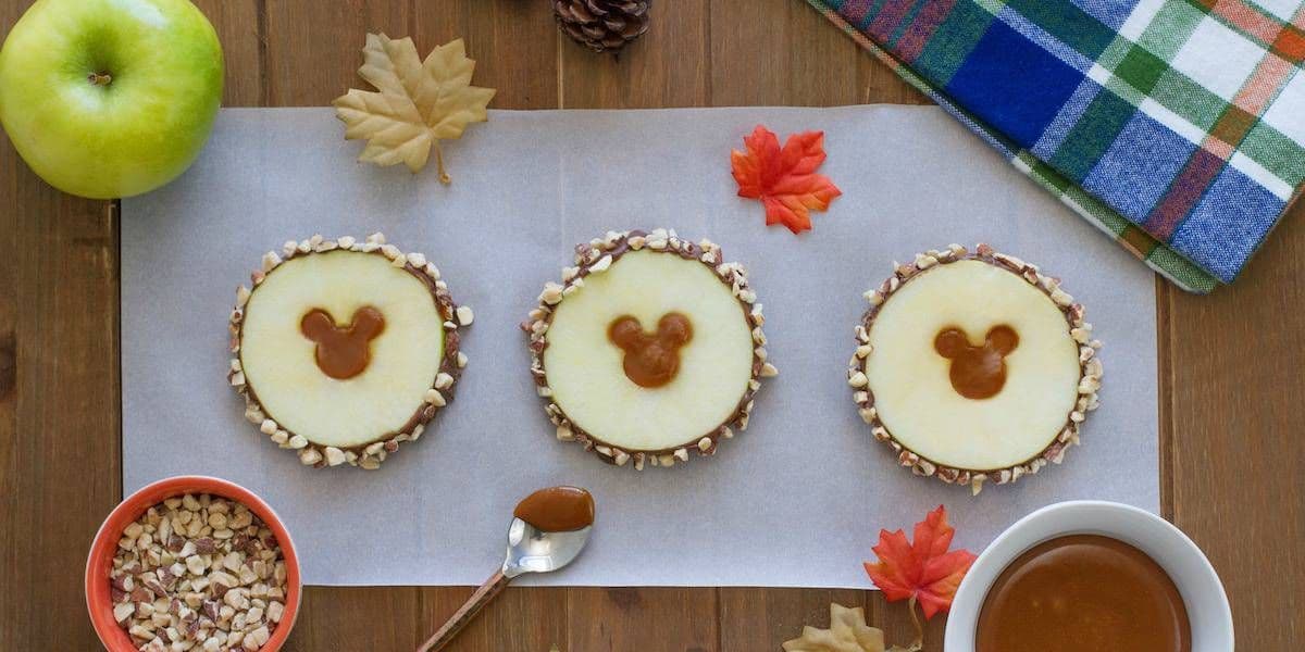 Three apple slices with caramel and nut coatings are arranged on parchment paper. Each slice has a Mickey Mouse-shaped caramel center. Surrounding the scene are leaves, an apple, nuts in a bowl, and a tartan-patterned napkin on a wooden surface.