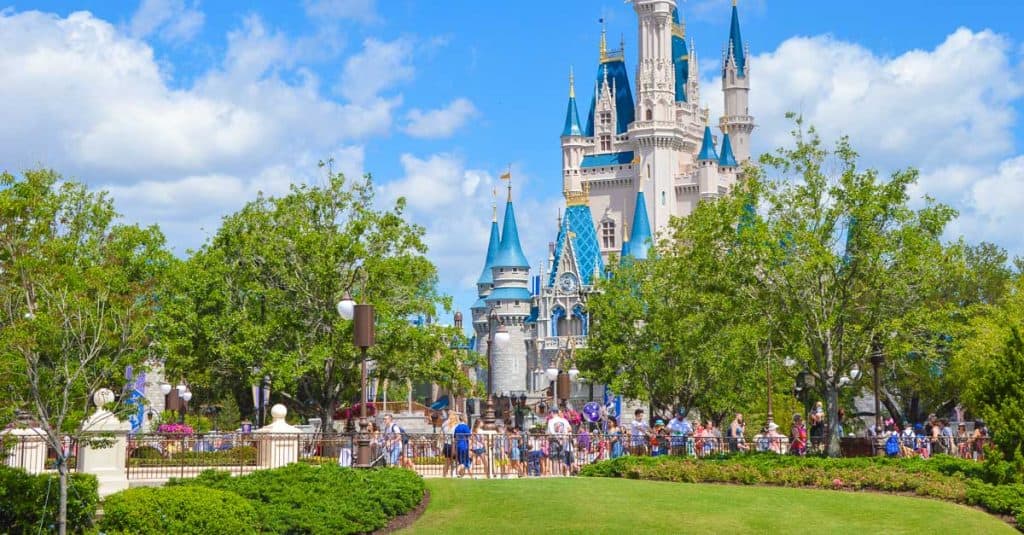 A large group of people walk and gather near Cinderella Castle at Disney World, surrounded by green trees and gardens under a bright, partly cloudy sky.