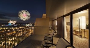 Resort balcony overlooking the city at night, with empty lounge chairs and fireworks lighting up the skyline like a theme park show.