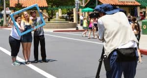 Three friends smile inside a blue photo frame as a family captures the excitement of Main Street U.S.A. on a bright day.