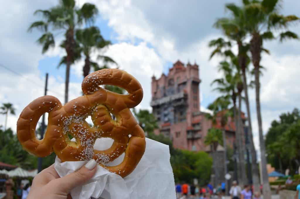 Mickey Mouse pretzel held up in front of Tower of Terror at Disney World, palm trees framing this classic theme park snack moment.