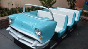 Sci-Fi Dine-In Theater's blue vintage car booth with white seats sits on gray pavement among lush greenery, ready for guests.