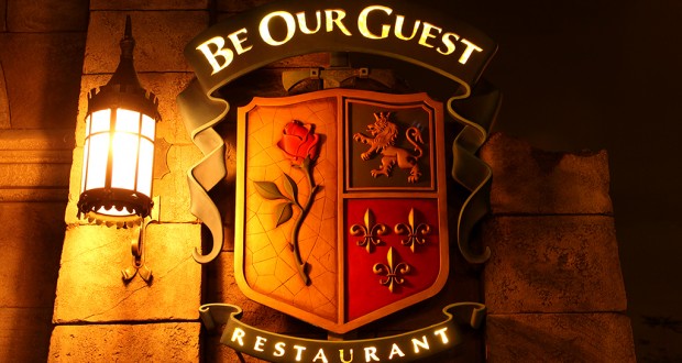 Be Our Guest Restaurant’s glowing sign, with rose, lion, fleur-de-lis, and castle shield icons, greets Disney World foodies.